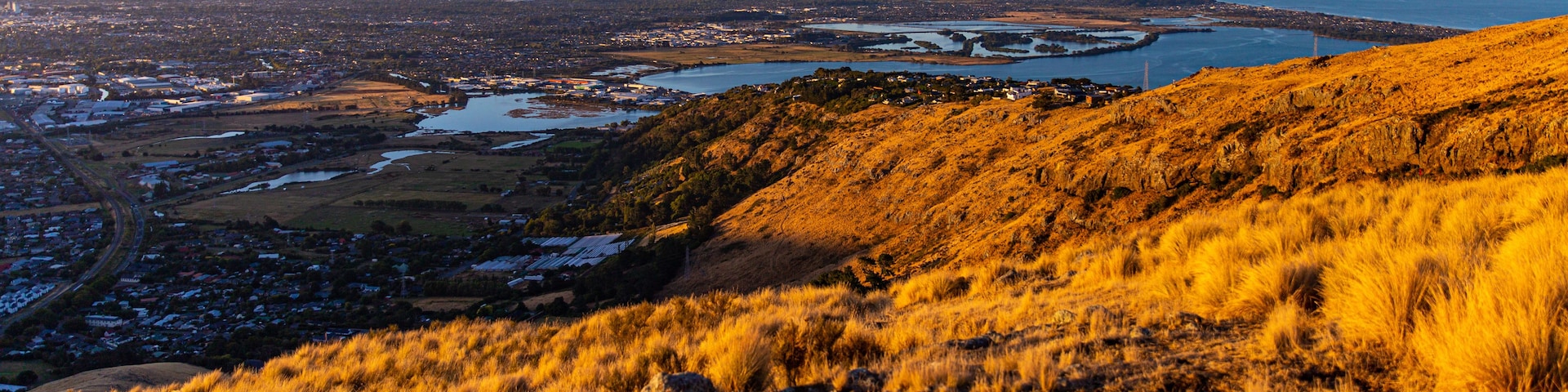 A coastal landscape with a hill in the foreground, a city in the middle ground, and a body of water in the background.