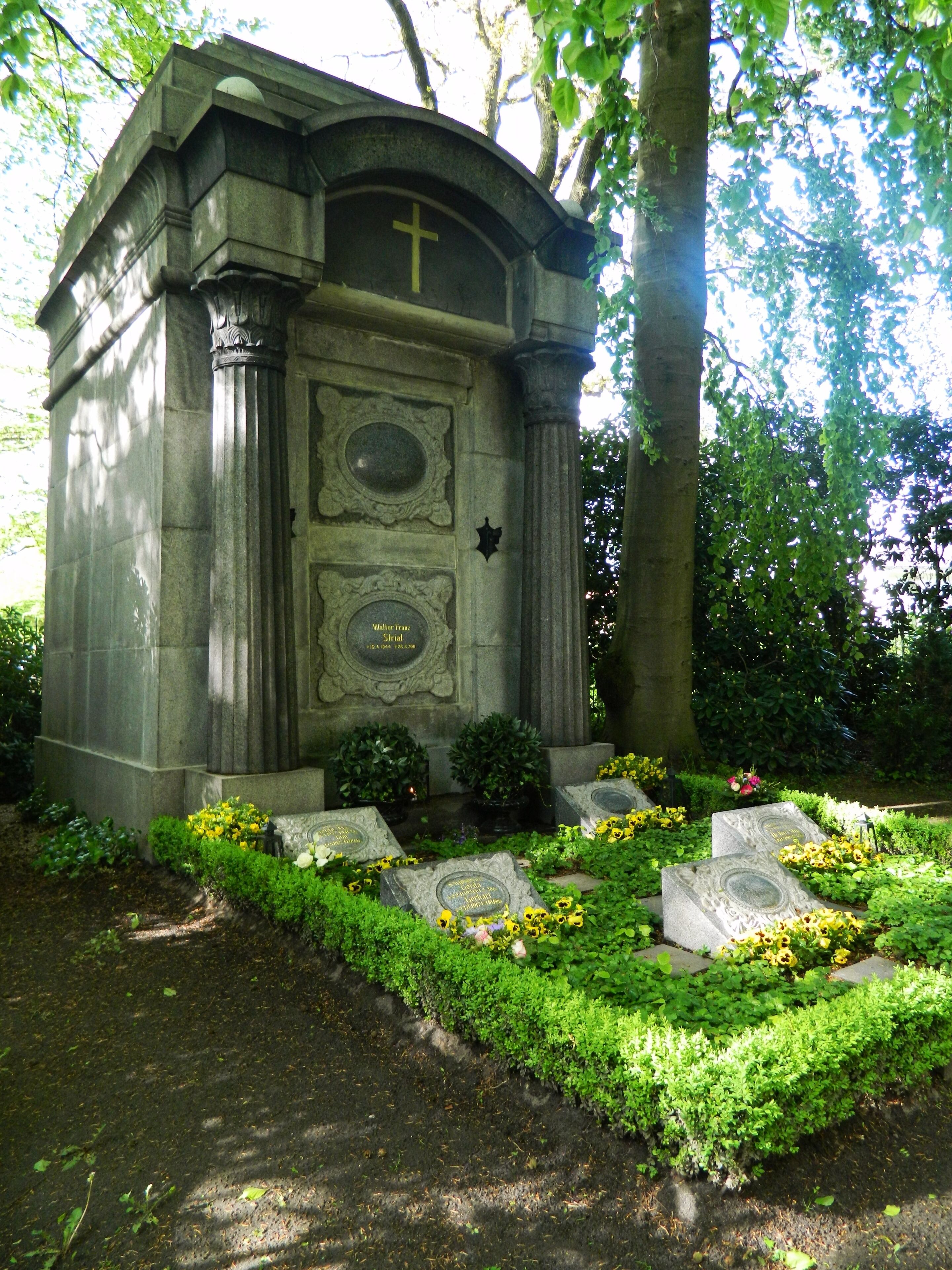 Monumento de piedra con una cruz, rodeado de follaje y flores.