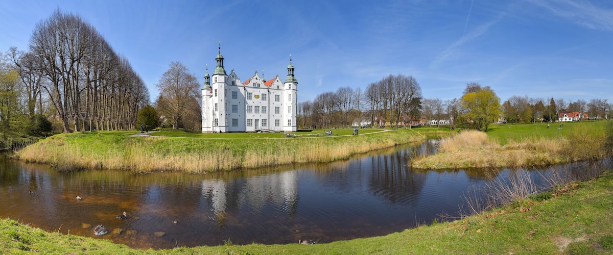 A large white castle with green spires, surrounded by a pond and trees.