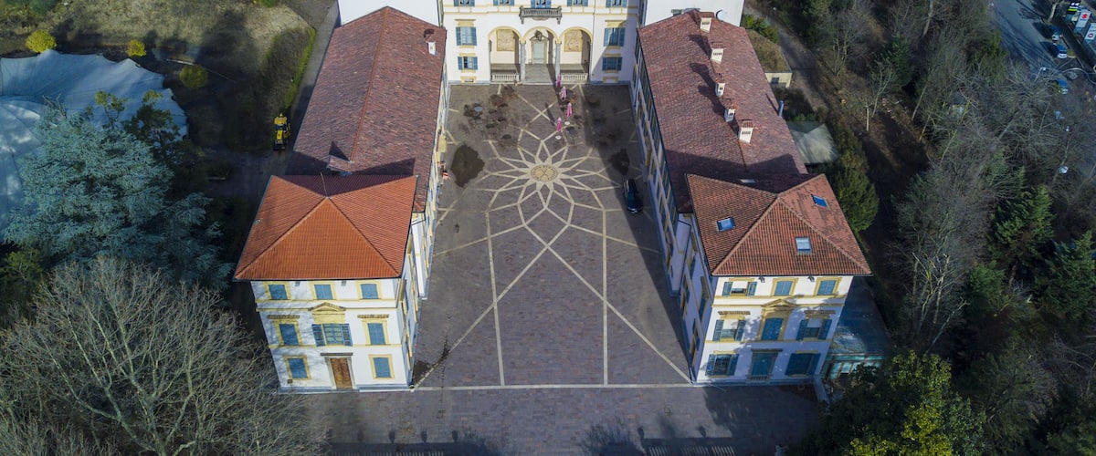 An aerial view of a large, symmetrical building with a central courtyard, surrounded by trees and parked cars.