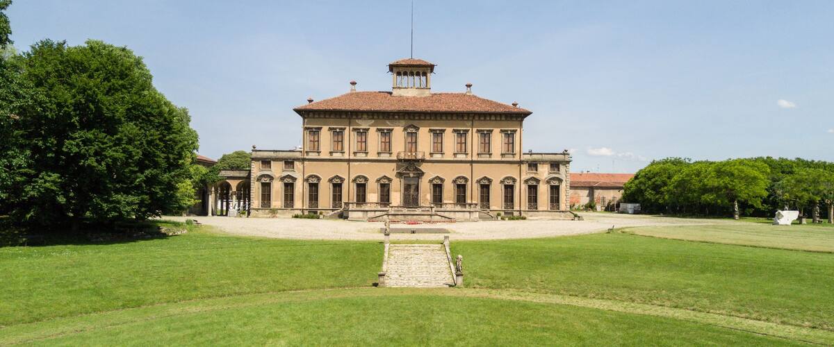 A large, historic building with a central staircase, surrounded by well-maintained lawns and trees.