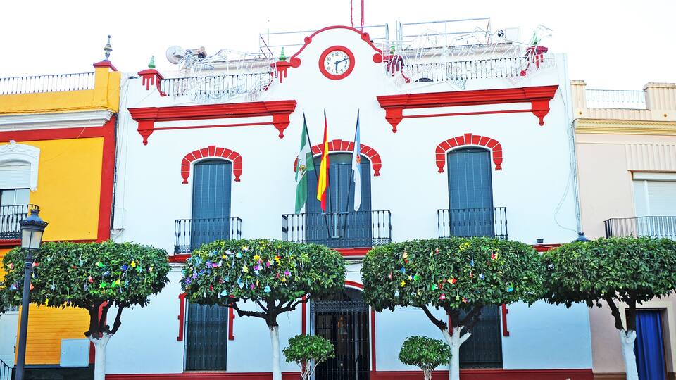 A white building with red trim and a clock. There are two trees in front of the building.