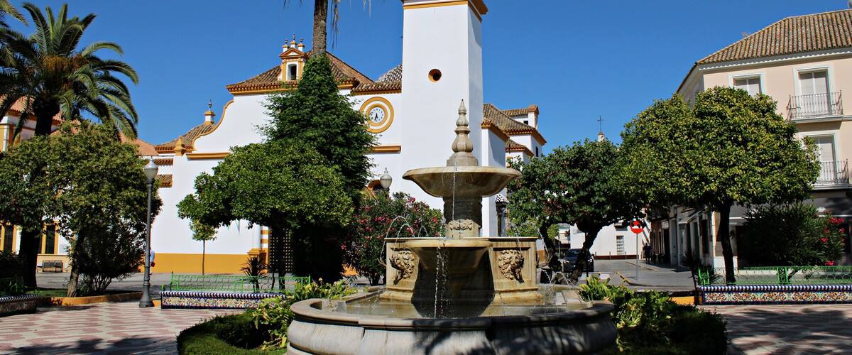 A fountain surrounded by greenery, a white church with a bell tower, and palm trees.