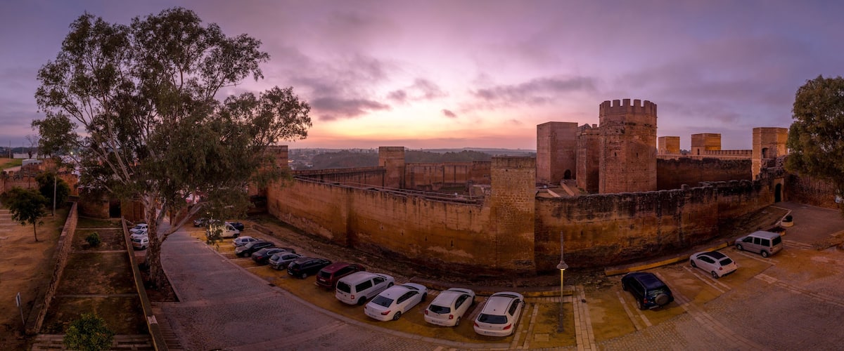 A historic castle with a moat, surrounded by trees and parked cars.