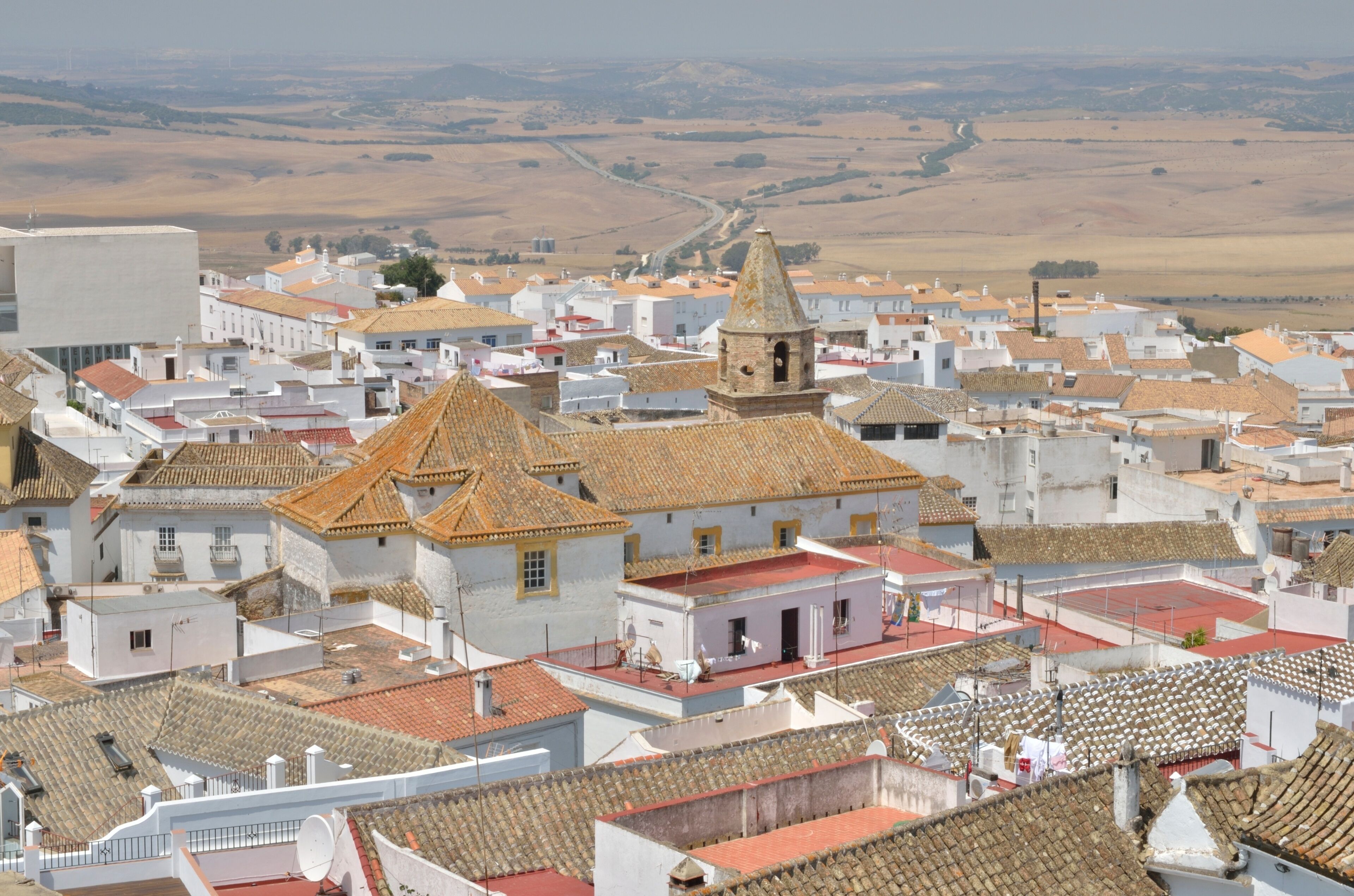 A town with white buildings and red-tiled roofs, a prominent church with a bell tower, and rolling hills in the background.