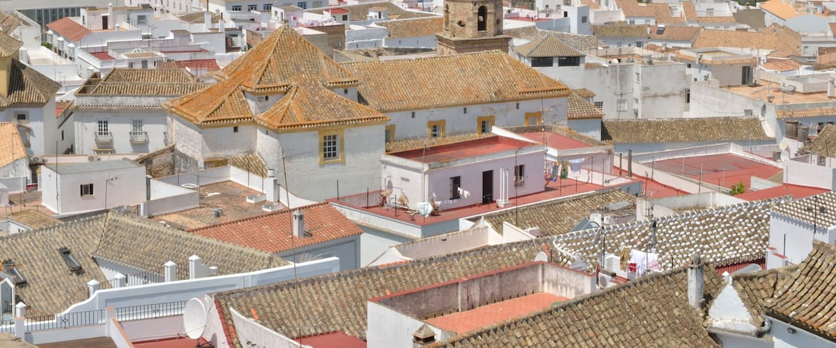 A town with white buildings and red-tiled roofs, a prominent church with a bell tower, and rolling hills in the background.