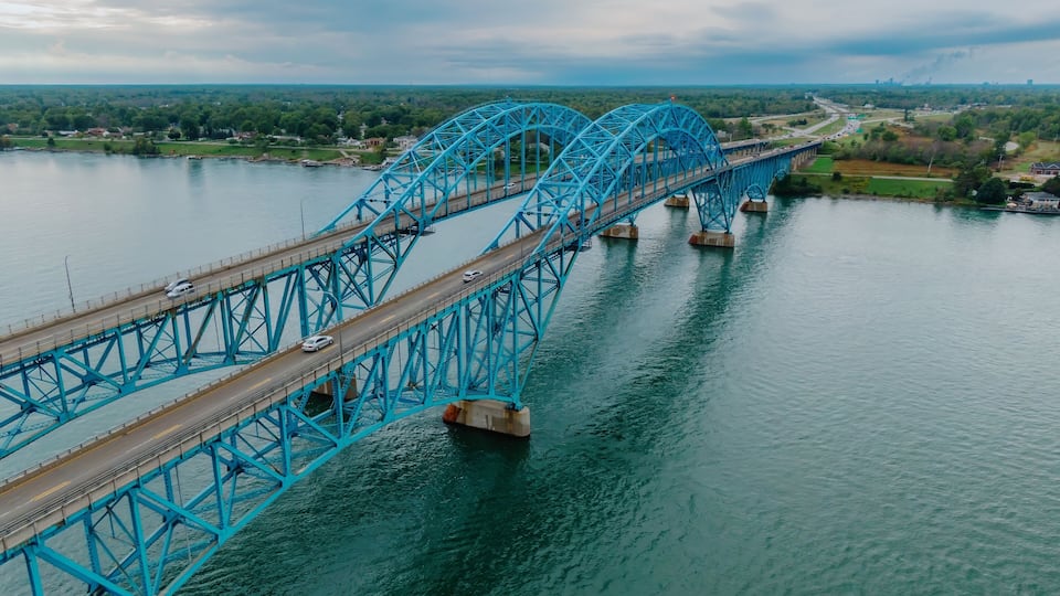 A large blue bridge spanning a river, surrounded by greenery and a cloudy sky.