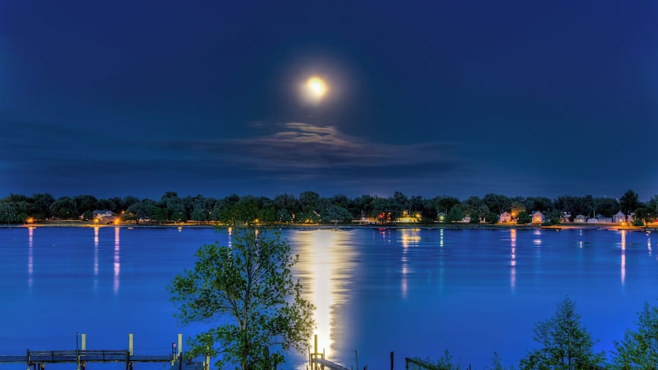 A calm lake at night with a full moon, dock, and houses along the shore.