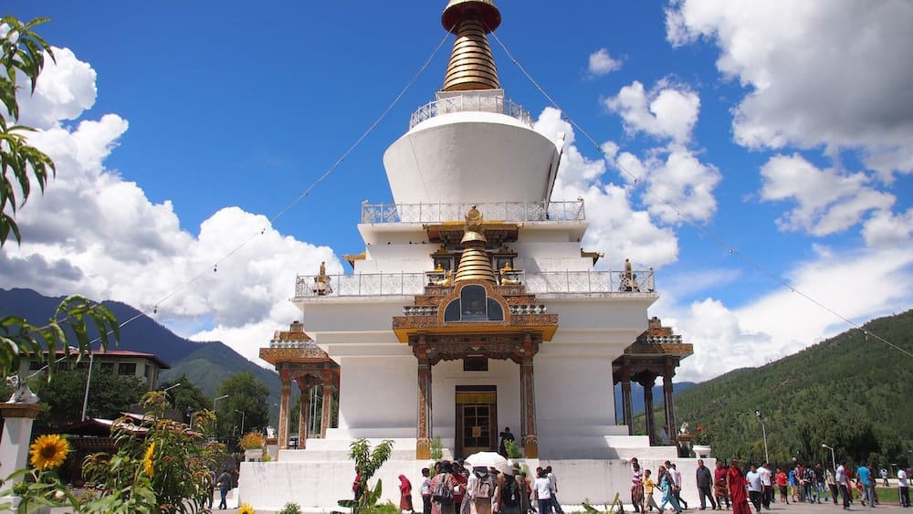 A white stupa with golden accents, surrounded by greenery and mountains.