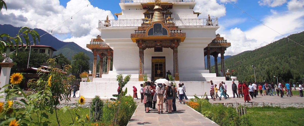 Um stupa branco com detalhes dourados, rodeado por vegetação e montanhas.