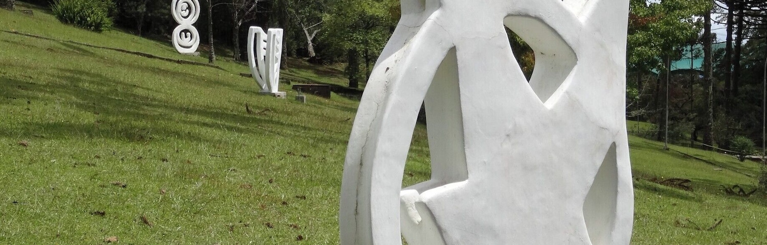 Escultura abstrata branca numa colina gramada, com árvores ao fundo.