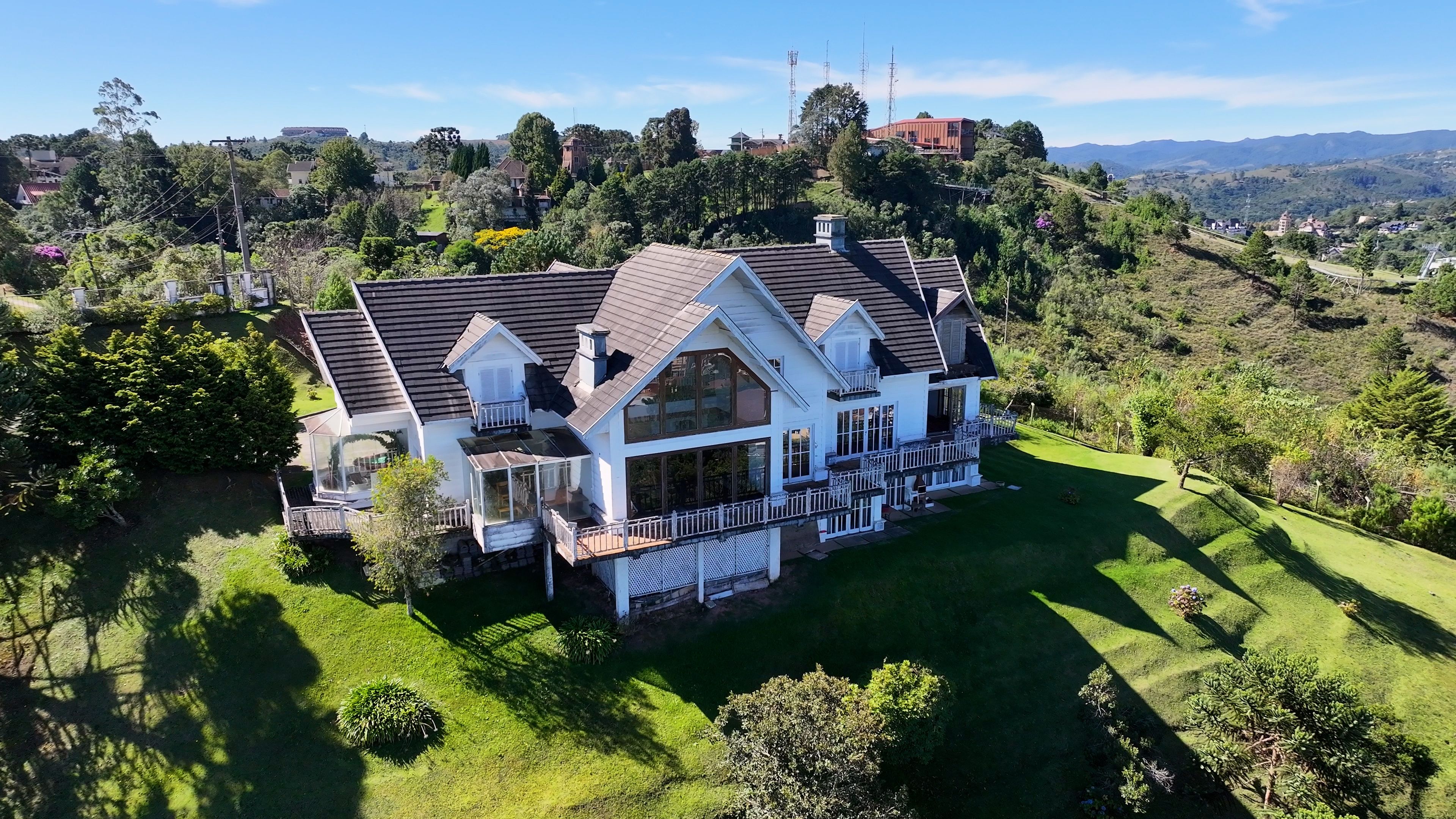 A large white house with a dark roof, surrounded by lush green grass and trees, set against a backdrop of rolling hills and distant mountains.