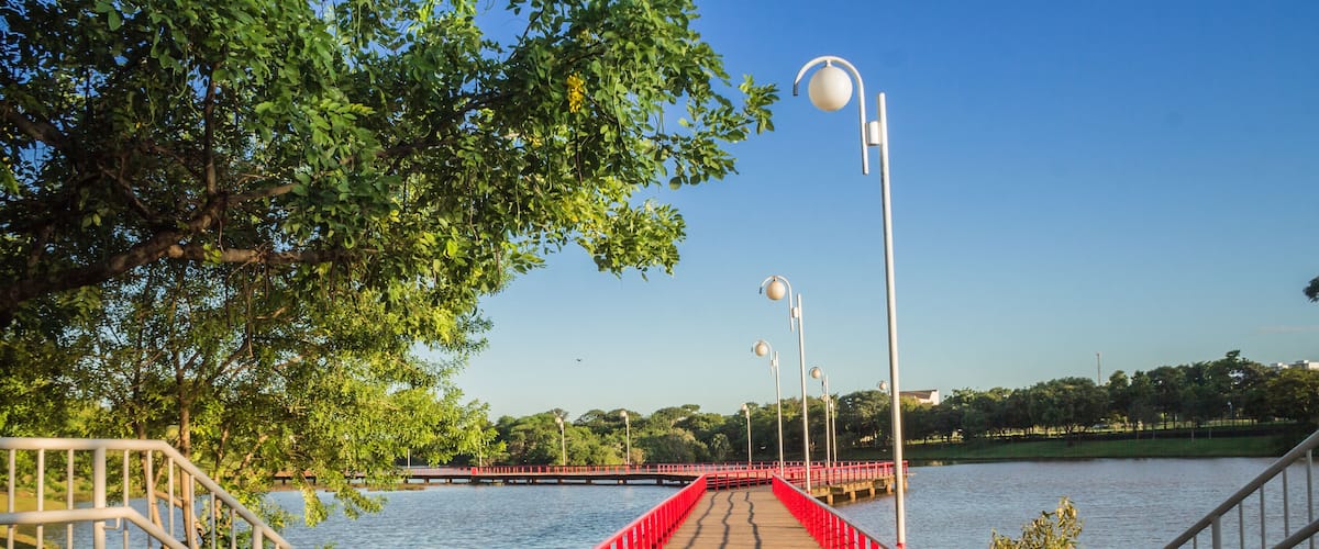 Un pont en bois avec des garde-corps rouges enjambant une étendue d’eau, entouré d’arbres et de lampadaires.