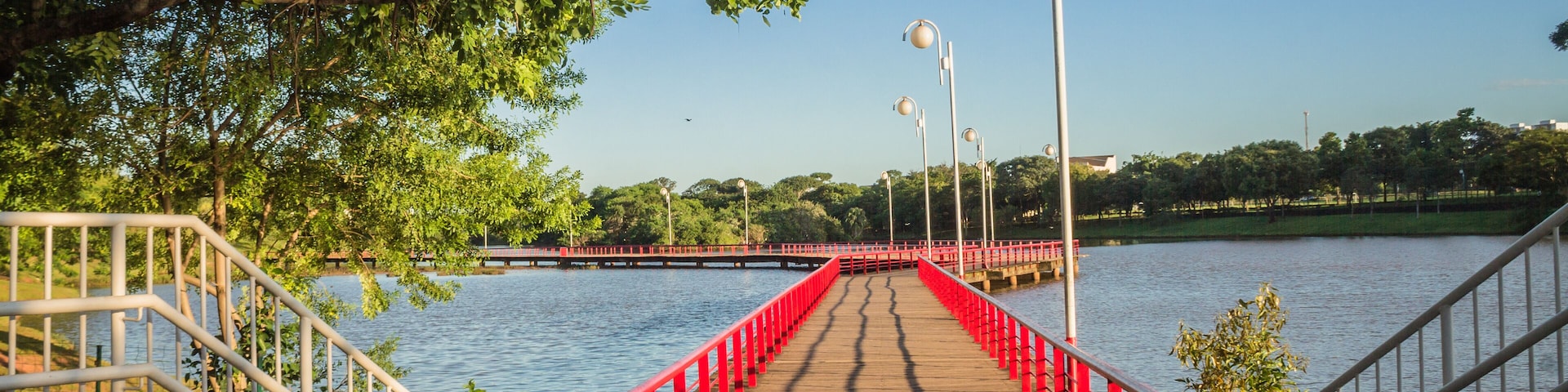 Uma ponte de madeira com grades vermelhas atravessa um corpo d'água, cercada por árvores e postes de luz.