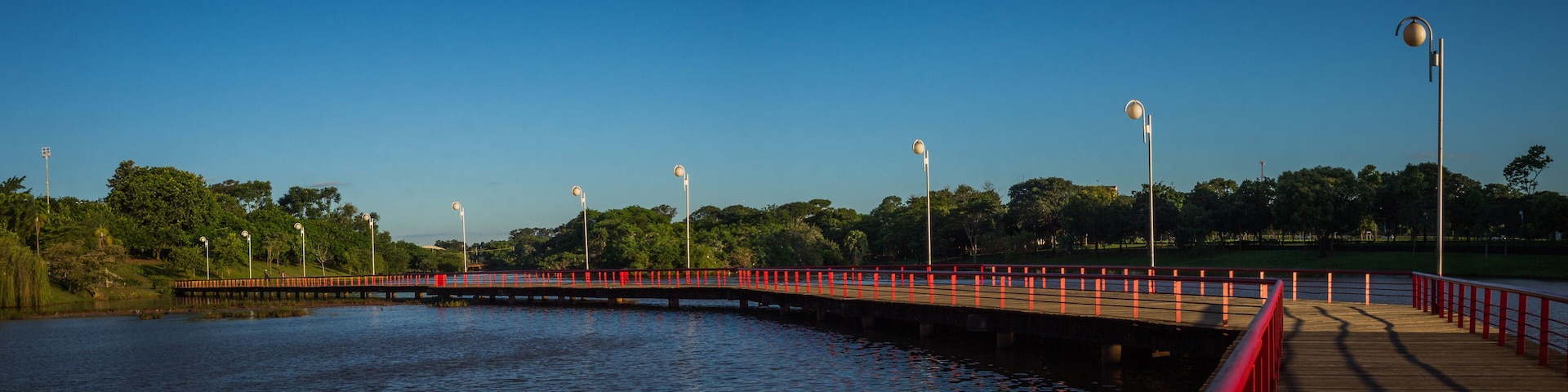 A wooden bridge with red railings over a calm body of water, surrounded by trees and street lamps.