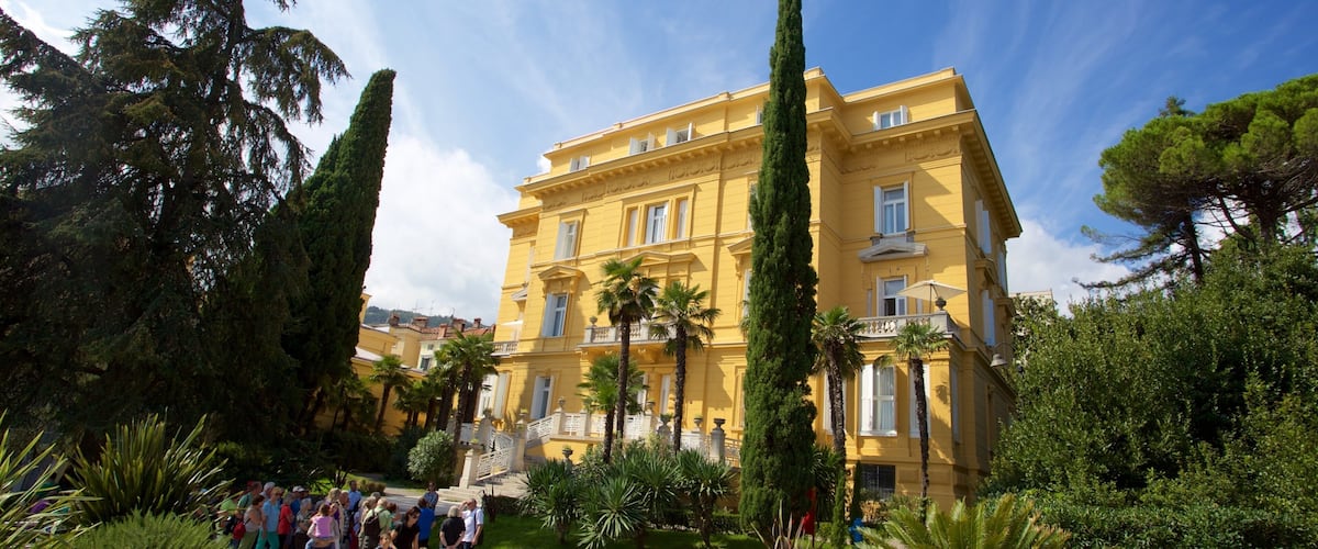 A yellow building with a balcony, surrounded by palm trees and cypress trees.