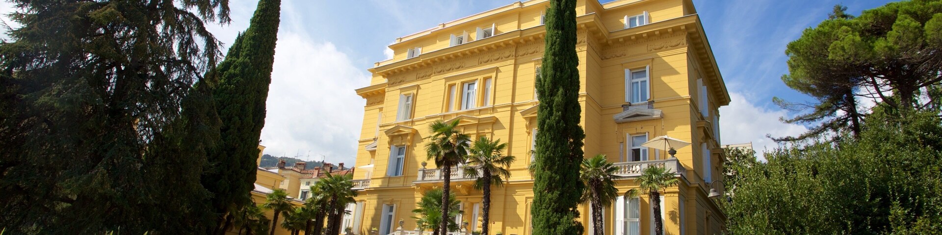 A yellow building with a balcony, surrounded by palm trees and cypress trees.