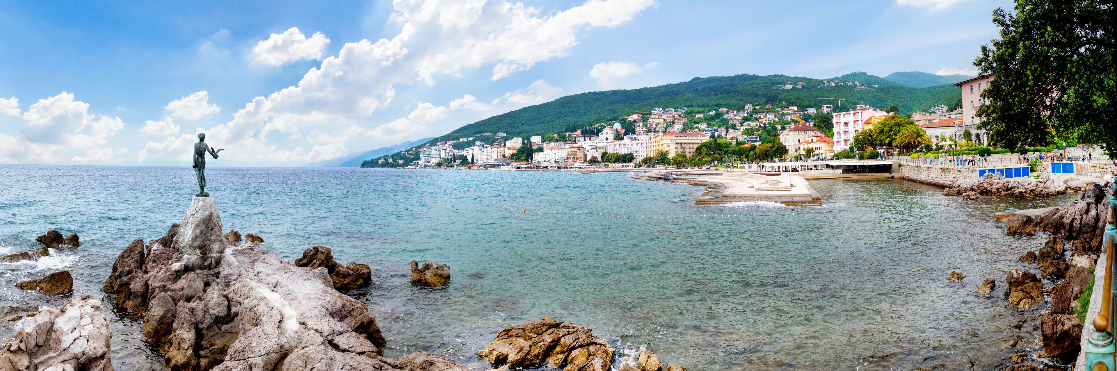 A coastal town with a statue on rocks, clear water, and buildings in the background.