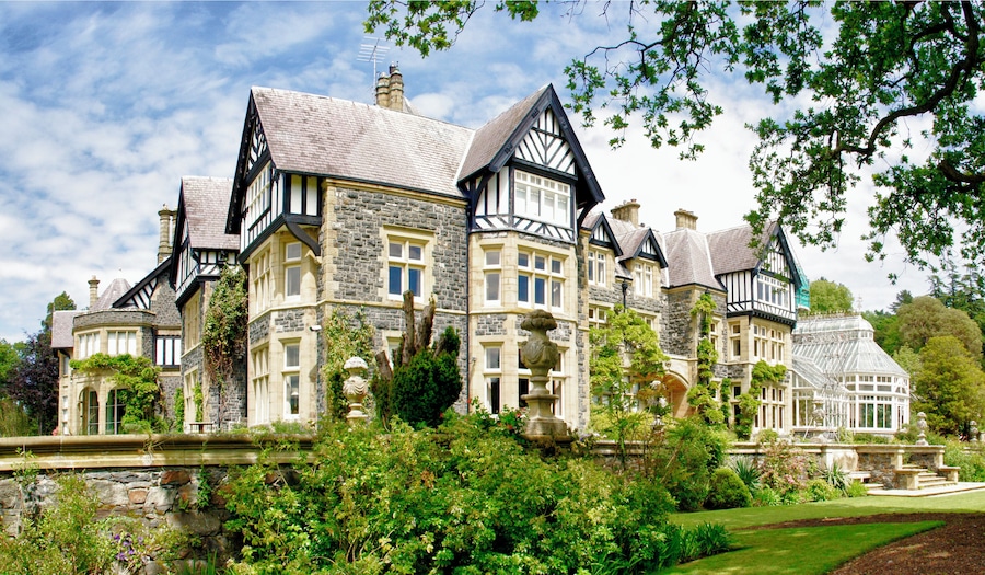 A large, historic house with stone and timber framing, surrounded by lush gardens and trees.