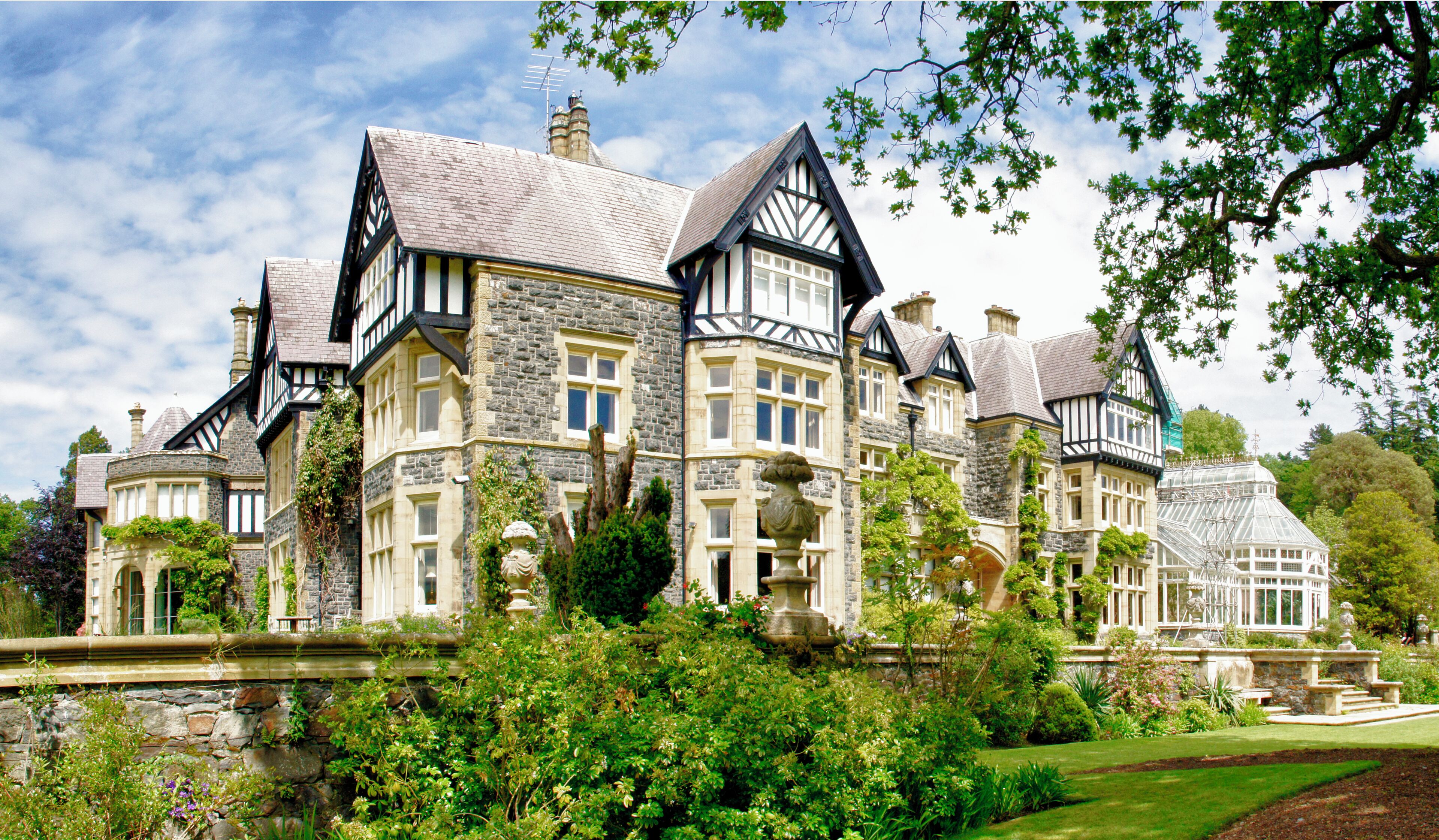 A large, historic house with stone and timber framing, surrounded by lush gardens and trees.