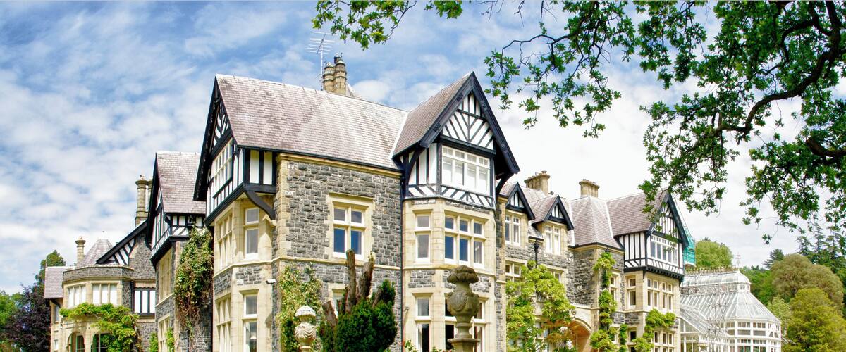 A large, historic house with stone and timber framing, surrounded by lush gardens and trees.