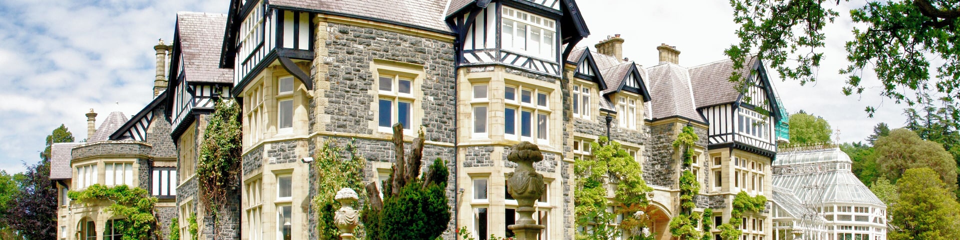 A large, historic house with stone and timber framing, surrounded by lush gardens and trees.