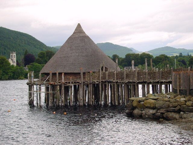 A thatched-roof structure on stilts by a lake, surrounded by greenery and hills.