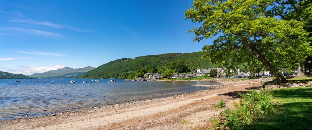 A lakeside scene with boats, a sandy beach, houses, and a forested hill.