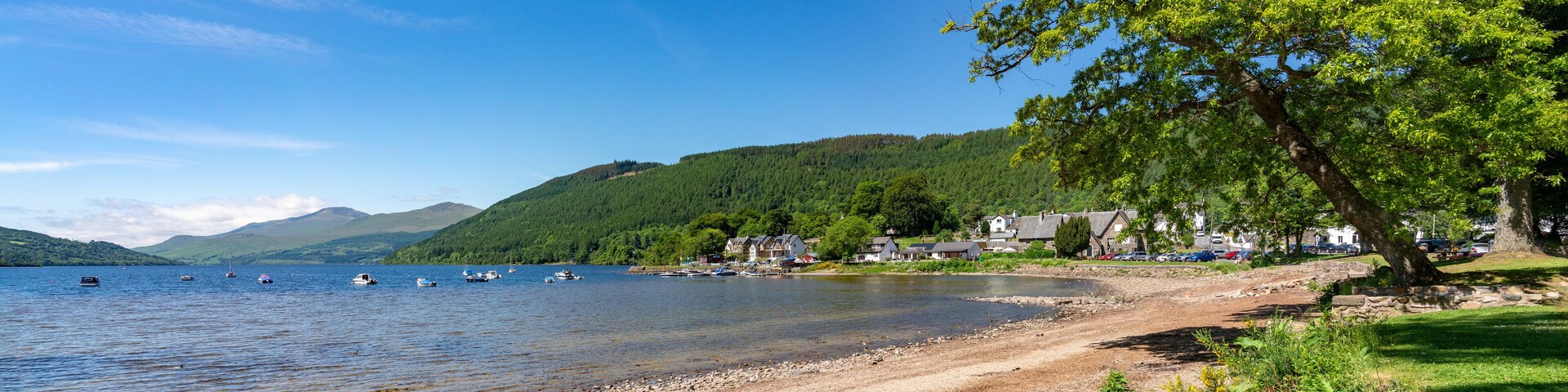 A lakeside scene with boats, a sandy beach, houses, and a forested hill.