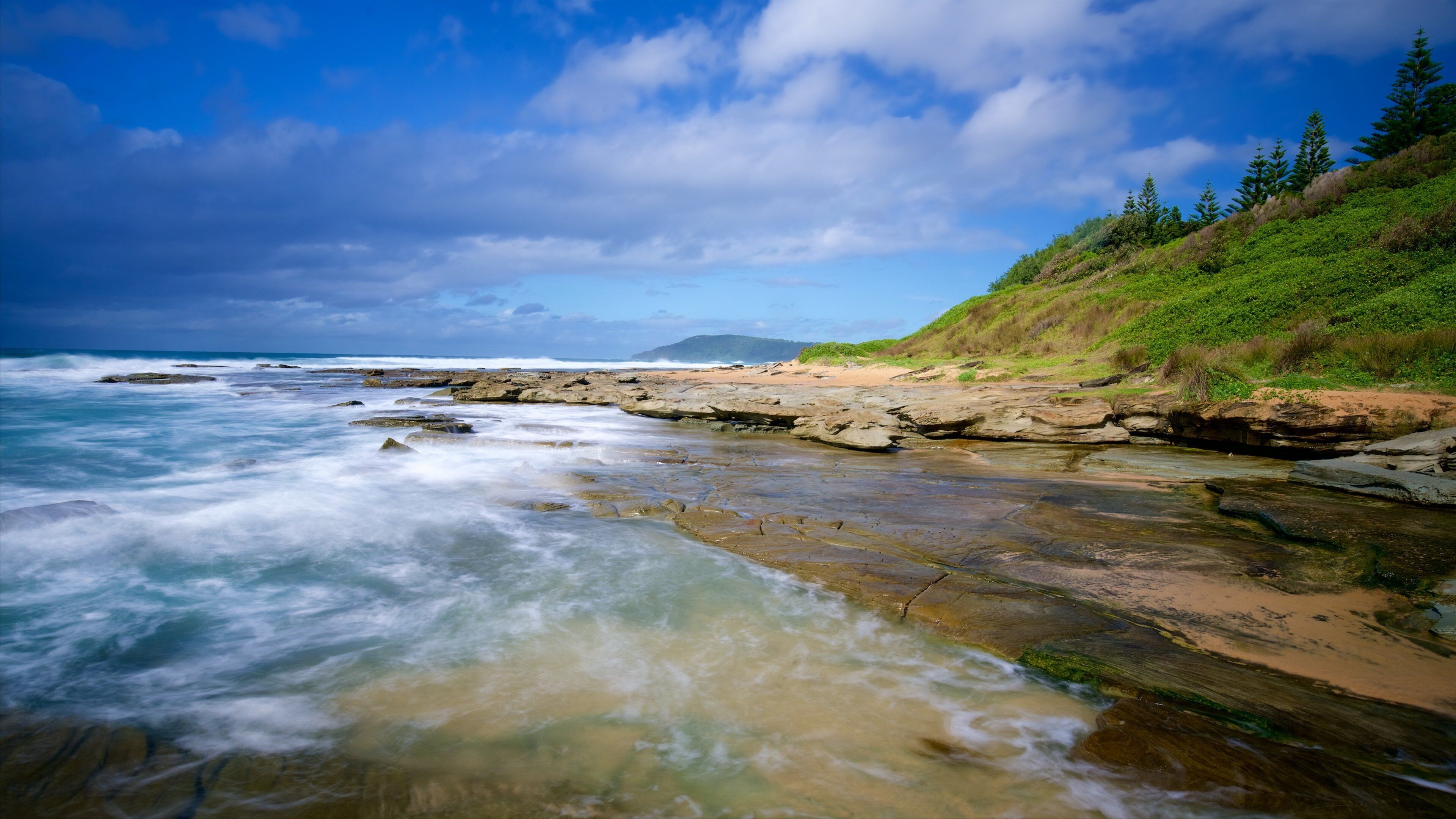 A rocky shoreline with waves crashing against the rocks, a sandy beach, and a grassy hillside with trees.