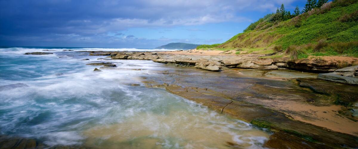 A rocky shoreline with waves crashing against the rocks, a sandy beach, and a grassy hillside with trees.