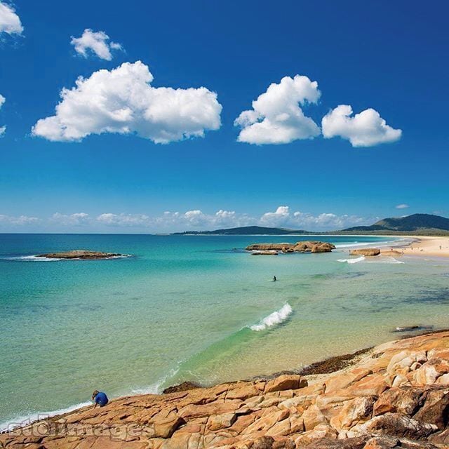A coastal landscape with clear turquoise waters, rocky shorelines, and distant mountains under a blue sky with clouds.