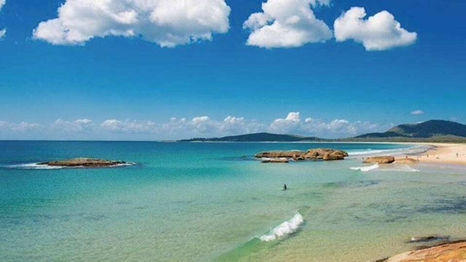A coastal landscape with clear turquoise waters, rocky shorelines, and distant mountains under a blue sky with clouds.