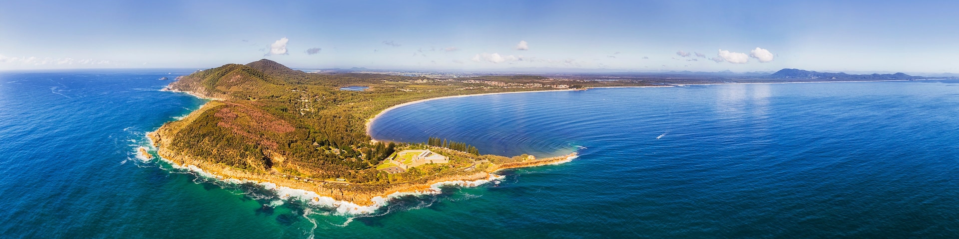 Vista aérea de uma ilha costeira com linha costeira curva, vegetação densa e mar azul cristalino.