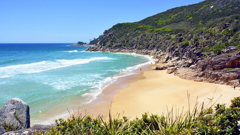 A coastal beach with clear turquoise waters, sandy shore, and a rocky hillside with greenery.