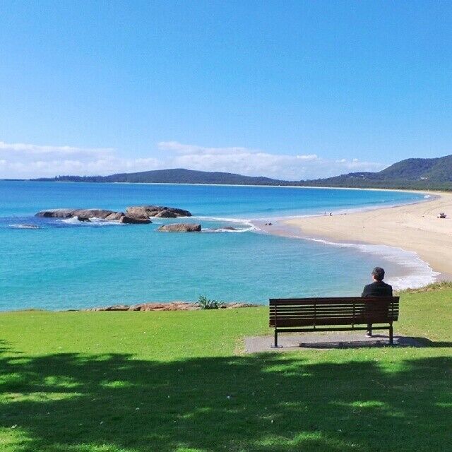 A beach with clear blue water, a sandy shore, and a person sitting on a bench.