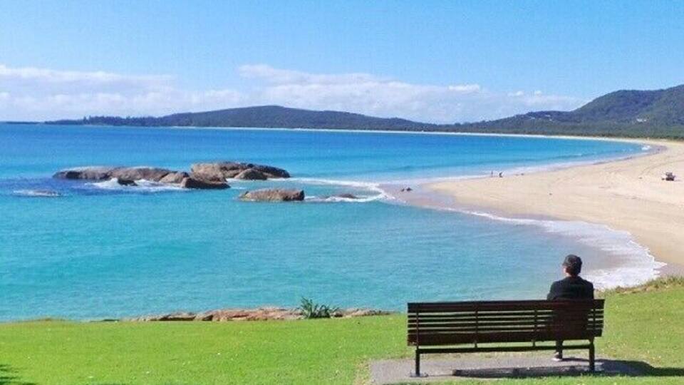 A beach with clear blue water, a sandy shore, and a person sitting on a bench.