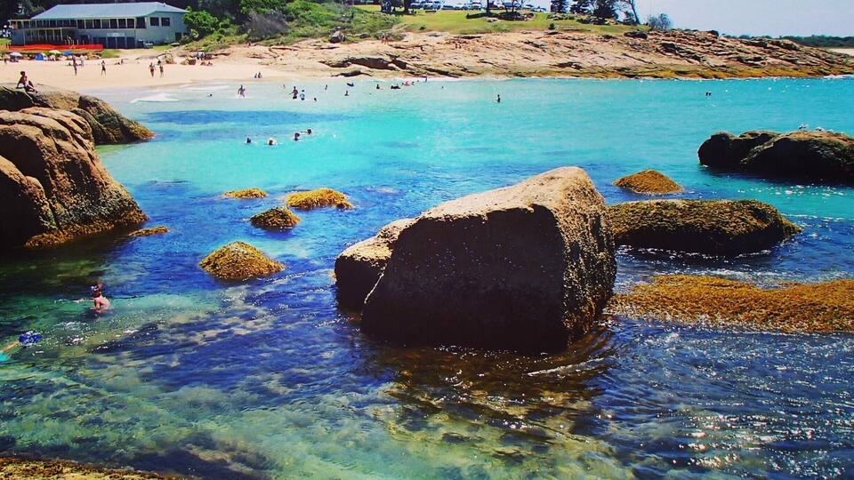 A coastal beach with clear turquoise water, large rocks, and a distant building.