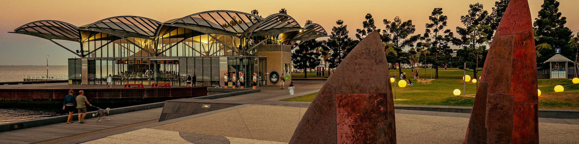 A waterfront promenade with modern architecture, including a glass building and a sculpture garden with large stone sculptures.