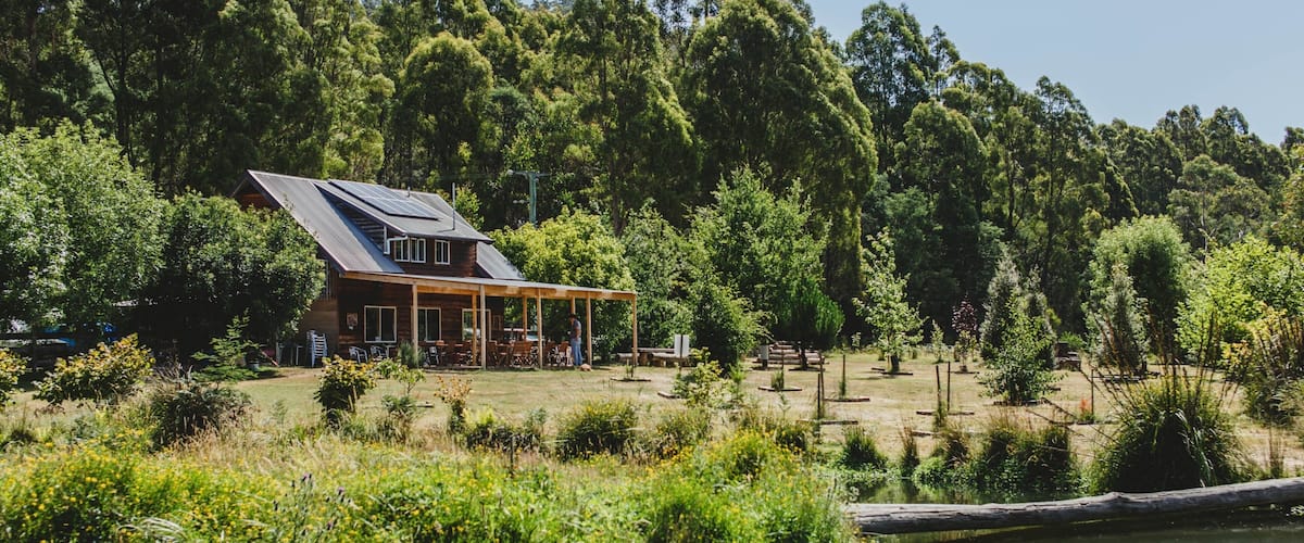 A wooden house with a sloped roof and a covered patio area, surrounded by dense forest and a body of water.