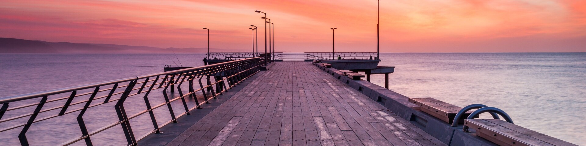 Un muelle de madera se adentra en un mar tranquilo bajo un cielo con una puesta de sol colorida.