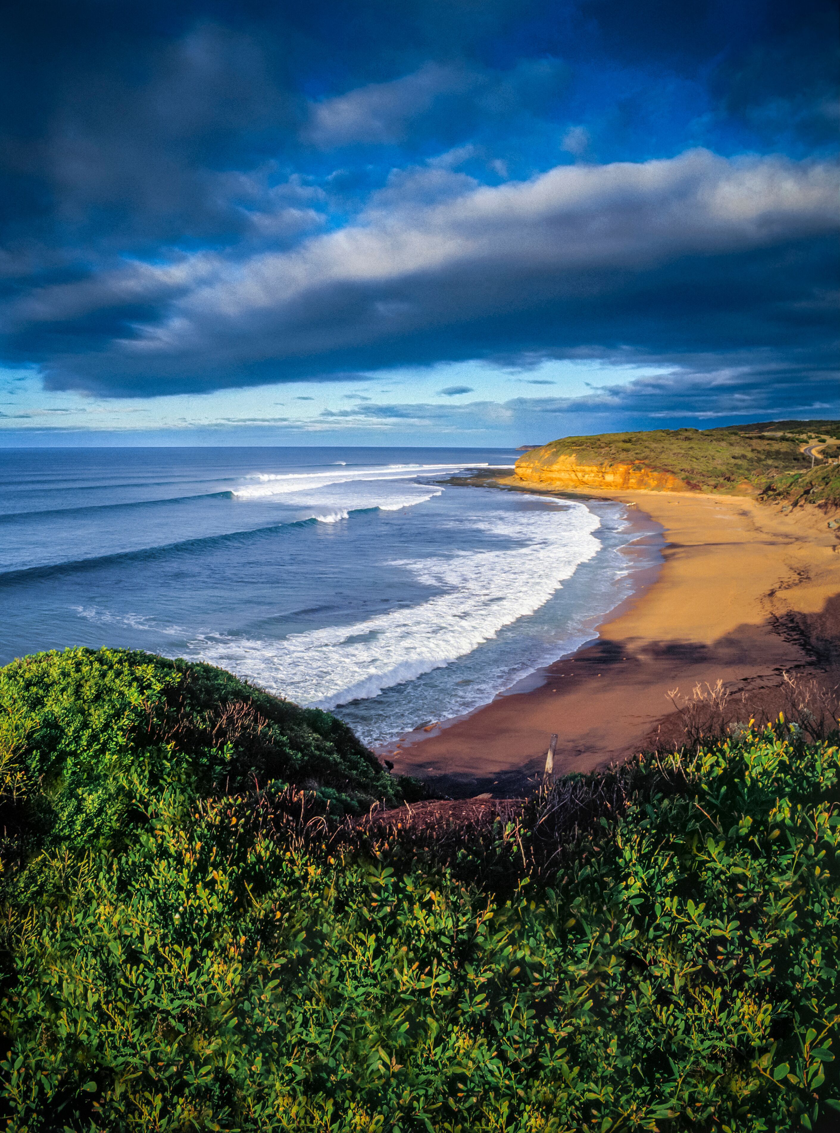 A coastal landscape with a sandy beach, ocean waves, and a cliff with vegetation.