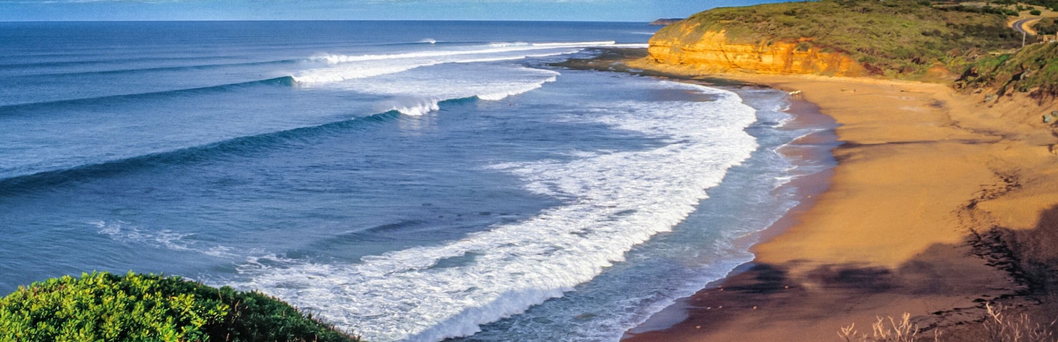 Un paisaje costero con una playa arenosa, olas oceánicas y un acantilado con vegetación.