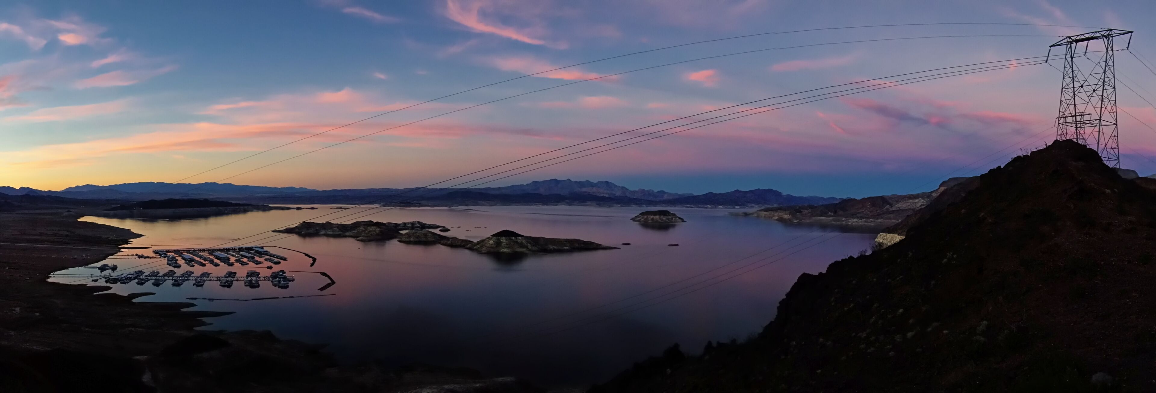 A calm lake with boats, a power line tower, and distant mountains during sunset.