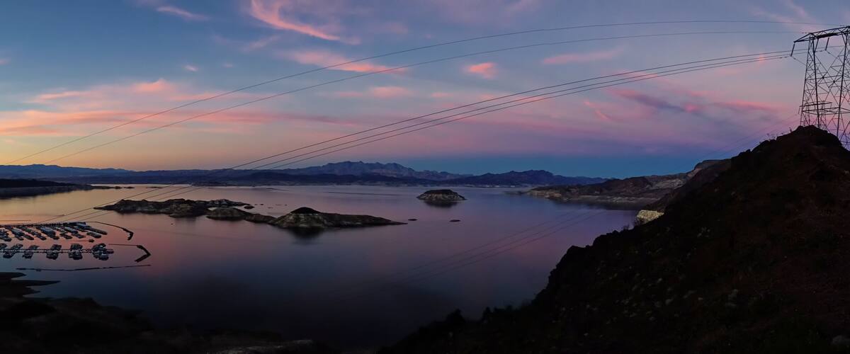 A calm lake with boats, a power line tower, and distant mountains during sunset.