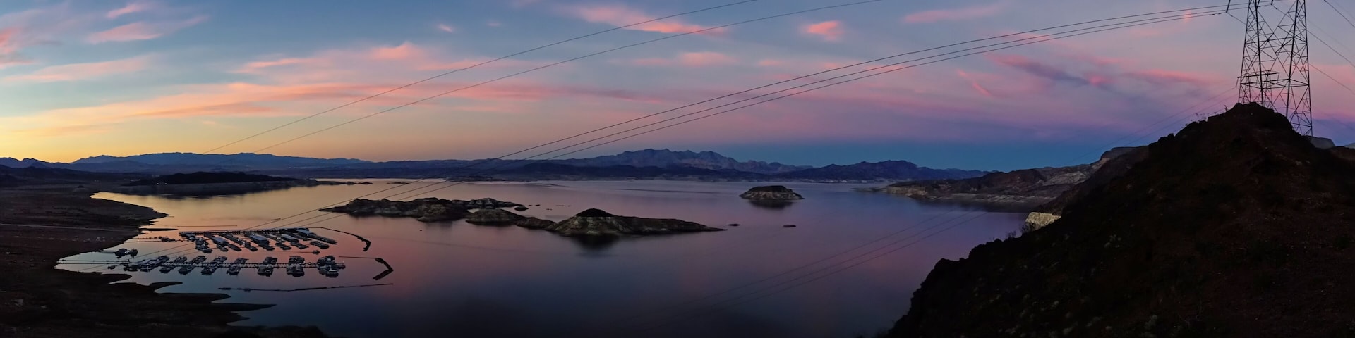A calm lake with boats, a power line tower, and distant mountains during sunset.