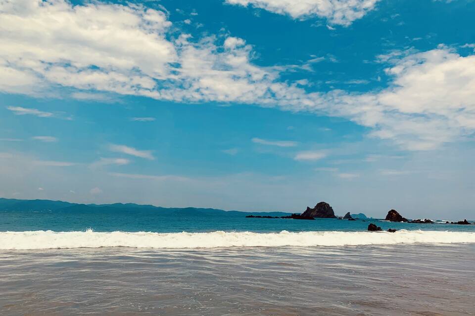 Una playa con olas, cielo despejado y montañas a lo lejos.