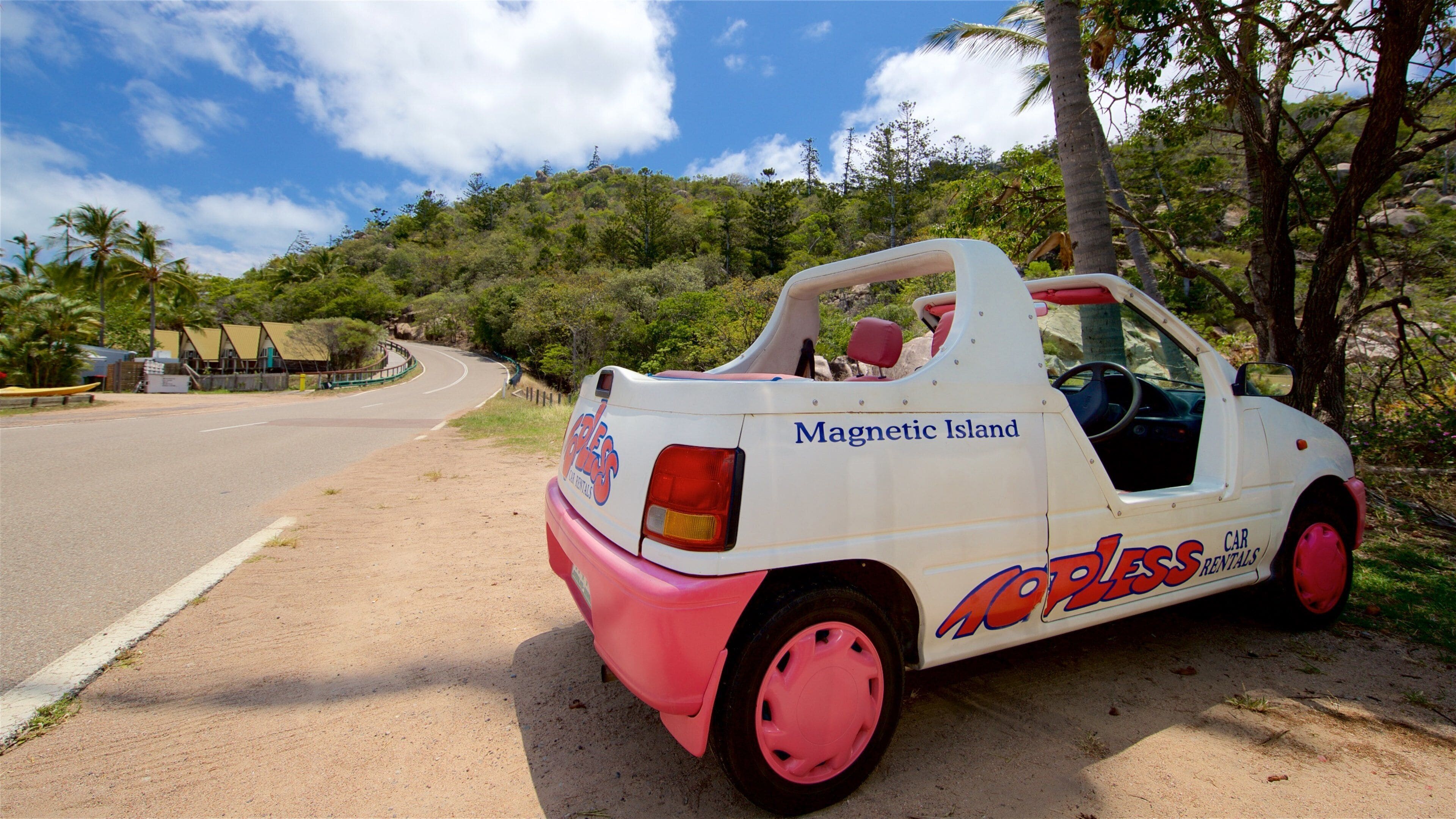 A white and pink car with "Carless Rentals" and "Magnetic Island" written on it, parked on a dirt road with a tropical landscape in the background.