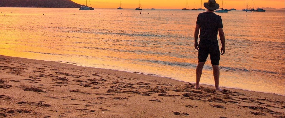 Een man staat op het strand bij zonsondergang, met zeilboten in de verte.
