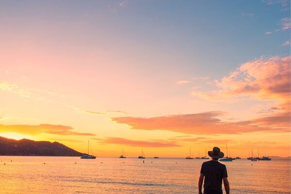 A man standing on a beach at sunset with sailboats in the distance.